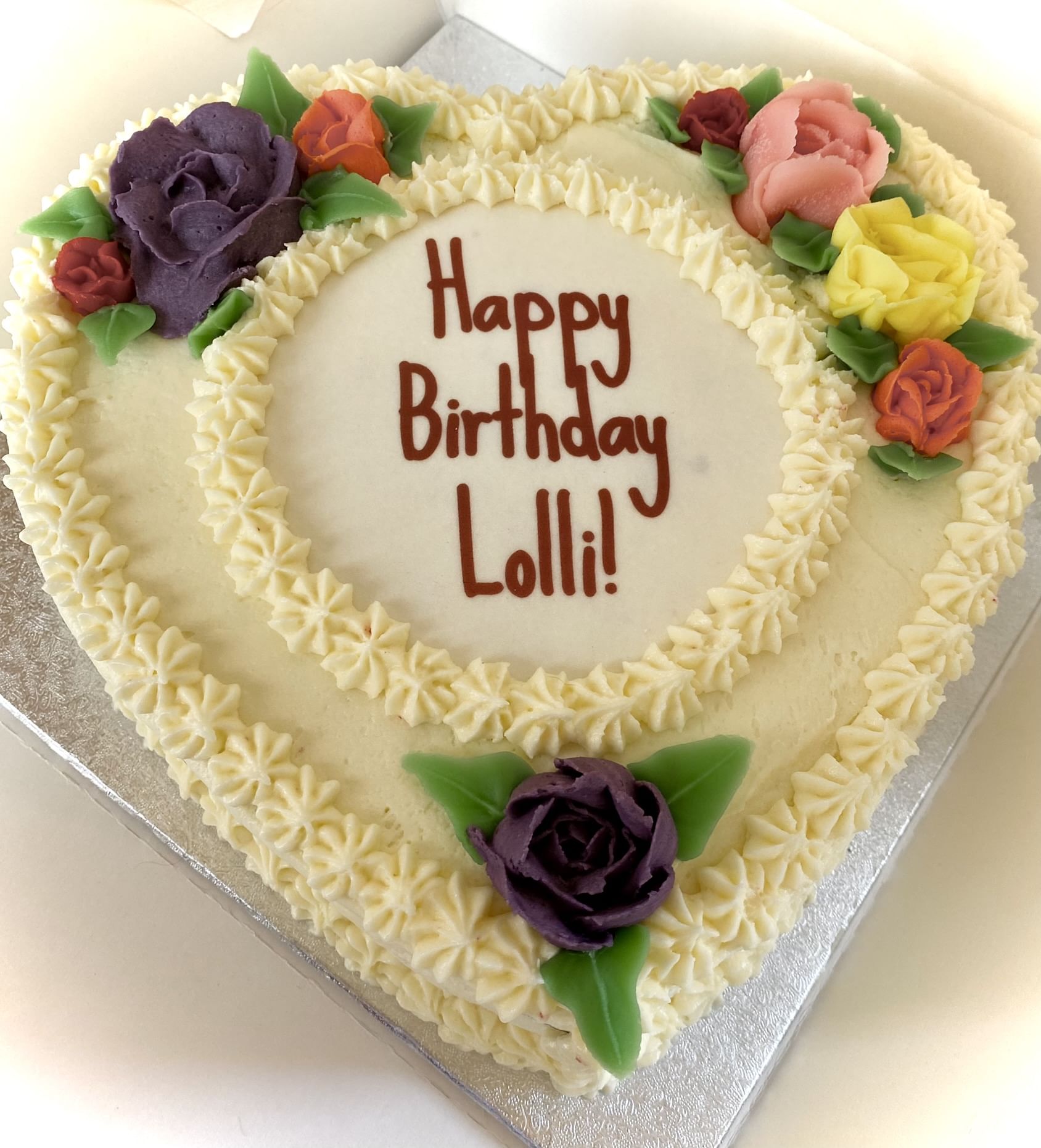 A heart-shaped cake with white frosting is decorated with colorful fondant roses in purple, red, yellow, and pink. This elegant design features two tiers, with the message 'Happy Birthday Lolli!' written in brown icing on the top tier—truly a sweet indulgence.